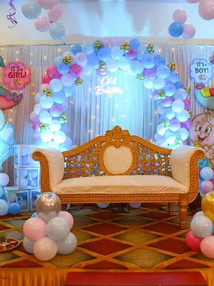 A wide shot of a baby shower stage decoration, featuring a blue and pink balloon arch and a beautiful couch for the parents-to-be.