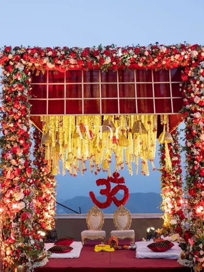 A traditional and divine mandap setup for a Silver Jubilee celebration. The structure is framed with a dense arch of red and white flowers, with a sacred 'Om' symbol at its heart.