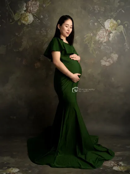 A classic maternity pose in a green gown against a vintage floral backdrop, creating a timeless and elegant portrait.