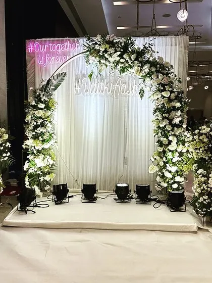 A modern photo backdrop with a circular arch covered in white flowers, complete with a custom hashtag and neon sign for a personal touch.