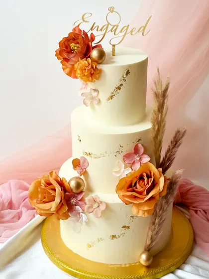 A magnificent three-tier engagement cake in off-white buttercream, decorated with gold leaf accents and a cascade of orange-toned roses and dried pampas grass.