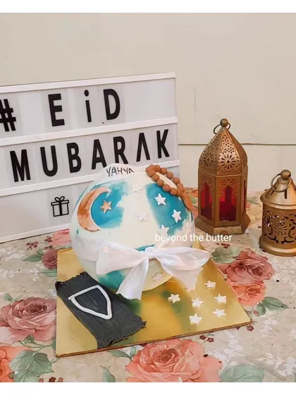 An Eid Mubarak cake designed as a sphere, decorated with a crescent moon and stars, and topped with a fondant prayer mat and tasbeeh.