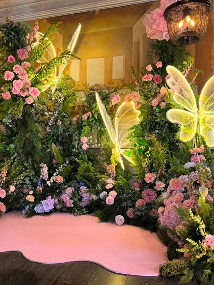 A close-up of the neon butterfly lights nestled within the lush greenery and pink flowers of the backdrop.