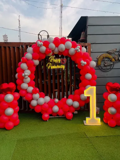 A vibrant first-anniversary party on a rooftop. This setup includes a circular balloon arch in red and silver, a neon sign, and a light-up number '1'.