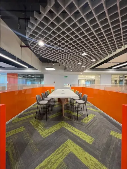 A long collaborative table set against a vibrant orange partition wall, designed for quick team huddles. The grid ceiling and patterned carpet define this zone within the larger workspace, demonstrating our use of design to guide function.