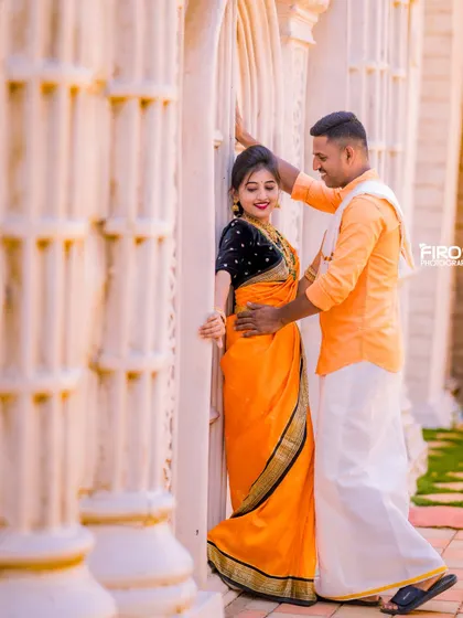 A romantic pose against a backdrop of white pillars. The bride's bright orange saree creates a stunning visual contrast.
