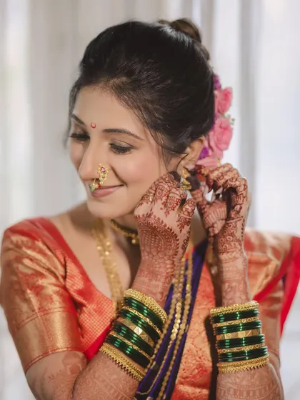 A close-up of the bride Aditi as she puts on her earrings. This shot highlights the detailed henna and the beautiful green bangles, essential parts of the Marathi bridal attire.