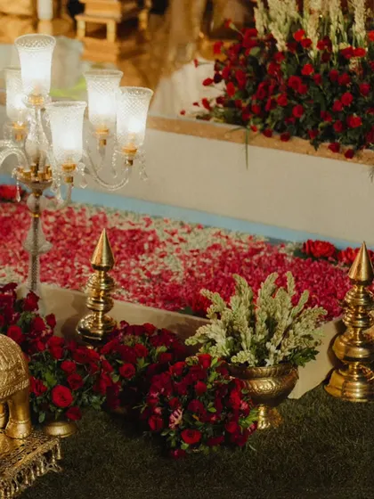 A close-up of the floral and brass decor lining the aisle. The path is covered in a carpet of red and pink rose petals, flanked by candelabras and traditional brass finials, creating a truly royal passage.