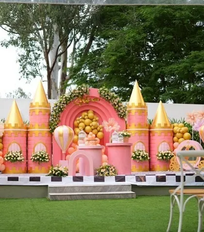 A wider view of Soha's pink and gold Disney-style castle, set up outdoors on a green lawn. The setup includes seating for guests, making it a perfect stage for a birthday ceremony.