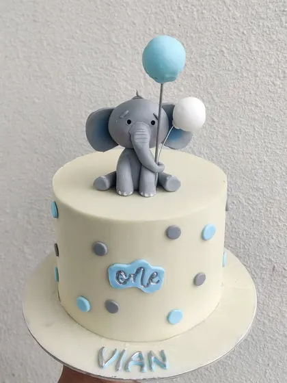 Sometimes, simple is the sweetest. This first birthday cake features an adorable, hand-sculpted elephant holding balloons, set against a clean white cake with blue and grey polka dots.