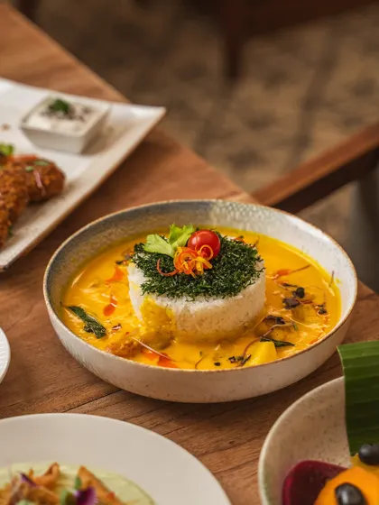 A beautifully presented curry dish with a mound of herb-topped rice. The natural lighting and wooden table create a rustic yet refined look for this fine-dining Indian plate.