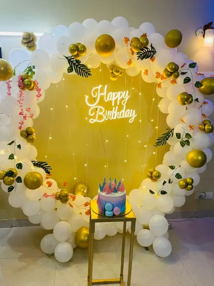 A simple and elegant birthday photo corner with a circular balloon arch in white and gold, a neon sign, and a plinth for the cake.