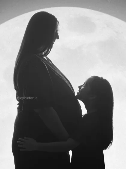 A powerful and creative black and white silhouette. This image tells a story of a growing family, with an older sibling looking up at her mother's baby bump against a full moon backdrop, symbolizing the new light coming into their lives.