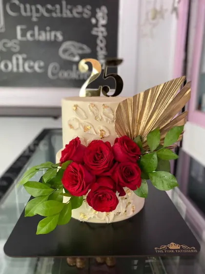 Celebrating 25 years with this elegant cake. The design includes a textured buttercream finish, a bouquet of fresh red roses, a modern gold palm leaf, and a "25" topper.