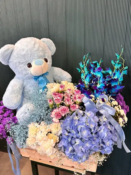 A different angle of the blue-themed teddy bear and flower crate. The mix of textures between the soft bear, delicate petals, and rustic wooden crate makes it a visually interesting gift.