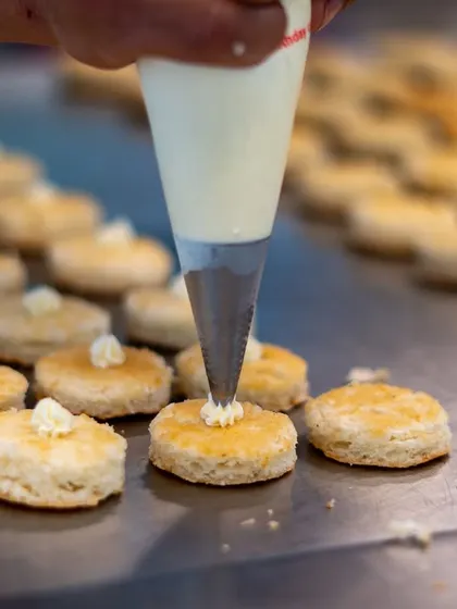 A close-up of our pastry team at work. A chef pipes delicate swirls of cream onto freshly baked scones, a small but crucial detail for an unforgettable high tea experience.