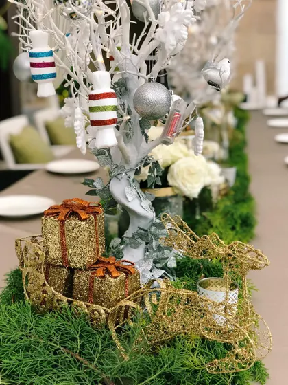 A close-up of a Christmas Eve dinner tablescape I designed. The centerpiece includes a white decorative tree with candy ornaments, surrounded by a lush green runner, glittery gold gift boxes, and a delicate wire reindeer, creating a festive and elegant holiday setting.