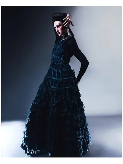 A full-length studio portrait featuring a model in a highly textured, dark blue gown. The dramatic pose and avant-garde styling showcase a high-fashion, conceptual aesthetic.