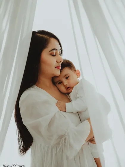 A mother holds her baby, who is looking over her shoulder with a curious expression. The soft, sheer curtains create a dreamy and ethereal effect.