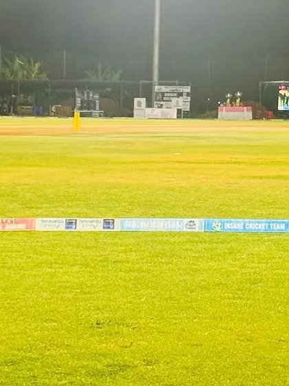 A view from behind the boundary rope during a night match, with the presentation area and scoreboard visible.
