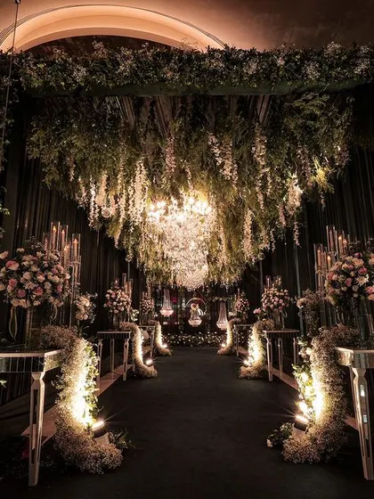 A dramatic and romantic entranceway, featuring a canopy of hanging florals and greenery, crystal chandeliers, and an aisle lined with illuminated floral arrangements.