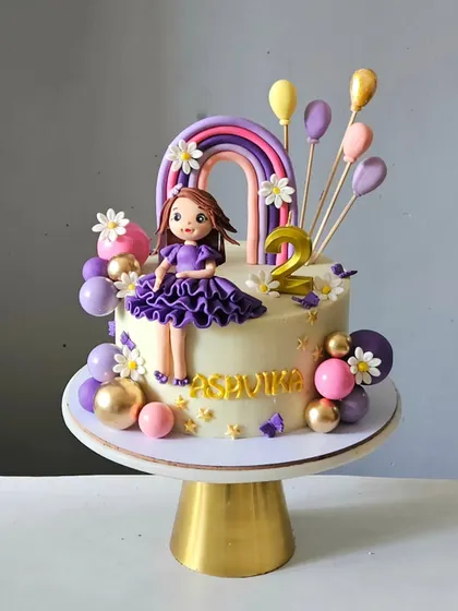 A beautiful dreamland cake for a second birthday. It features a custom girl figurine in a purple dress, a pastel rainbow, and balloon accents, all handcrafted to create a whimsical and celebratory design.