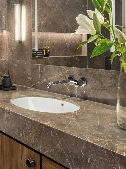 This bathroom in the West Coast Residence features a darker, moodier palette with a rich brown marble. Backlighting behind the mirror niche creates a soft, ambient glow.