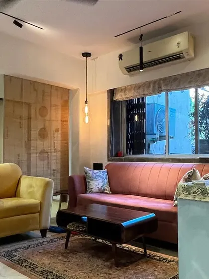 An eclectic living room with a mix of furniture styles, including a pink mid-century modern sofa and a yellow armchair. The lighting combines track lights with a decorative pendant light.