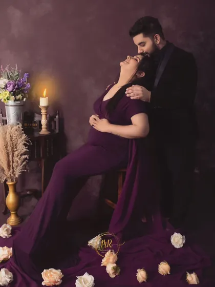 A romantic, candlelit portrait of a couple. The partner kisses his wife's forehead as she leans back, surrounded by soft light and scattered rose petals.