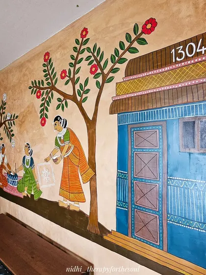 Another angle of the village scene, highlighting the vibrant blue door and the woman drawing a rangoli, showcasing the storytelling aspect of Cheriyal folk painting.