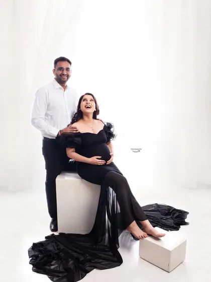 A joyful and relaxed couple portrait. The mom-to-be is seated in a beautiful black gown while her partner stands behind her, sharing a happy moment in a bright, high-key setting.