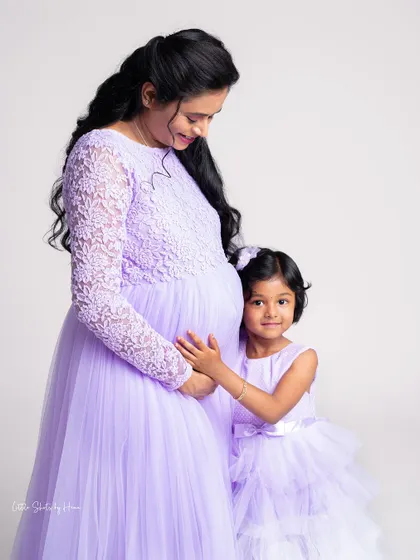 A mother and daughter share a special moment, both dressed in matching lavender gowns. The daughter gently touches her mother's baby bump, excited to become a big sister.