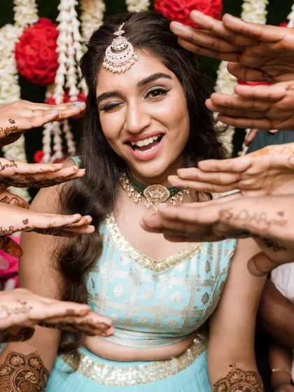 Fun and candid moments from a mehendi ceremony. The bride's makeup is fresh and playful, with a winged liner and glowing skin, perfect for celebrating with her loved ones. The hairstyle is a soft half-updo with pretty accessories.