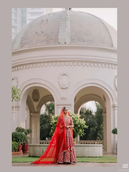 A full-length portrait of a bride standing before a classic white rotunda, her red lehenga providing a vibrant contrast to the elegant backdrop.