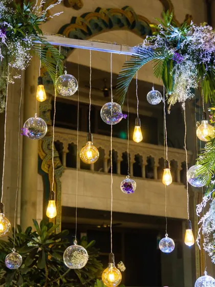 This decor detail shows a mix of vintage-style Edison bulbs and clear glass orbs hanging from a lush green floral arrangement, providing warm and stylish lighting for the party.