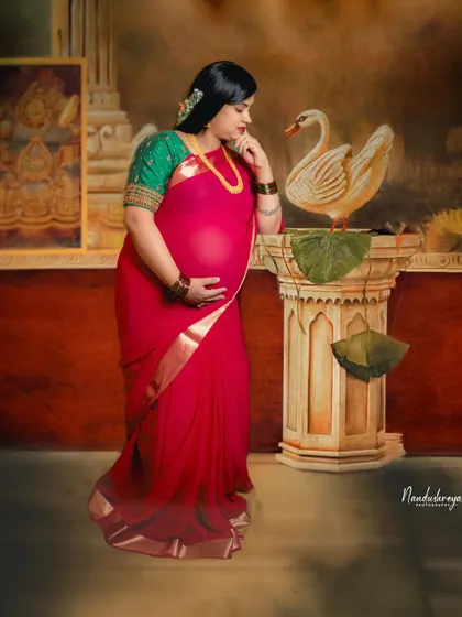 A beautiful studio portrait in a red saree against a painted backdrop. The swan prop adds a touch of grace and elegance to this traditional maternity photo.