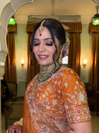A smiling bride, ready for her wedding. Her hair is styled in a beautiful floral bun, with soft tendrils framing her face, complementing her glowing makeup and heavy jewelry.