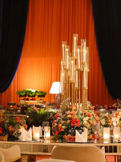 The dining setup for the poker night, with tables surrounded by lush arrangements of orange and red flowers. The tall candelabras add a touch of classic elegance to the modern, vibrant setting.