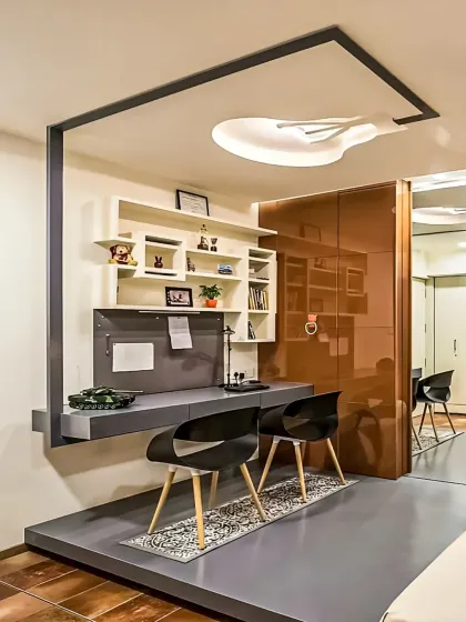 We designed this unique home office nook on a raised platform. The floating grey desk is framed by a black metal structure, and the wall-mounted shelves provide ample storage, creating a distinct and modern workspace within a larger room.