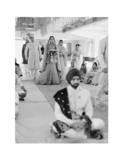 A black and white photograph capturing the bride's solemn and graceful entrance during a Sikh wedding ceremony.