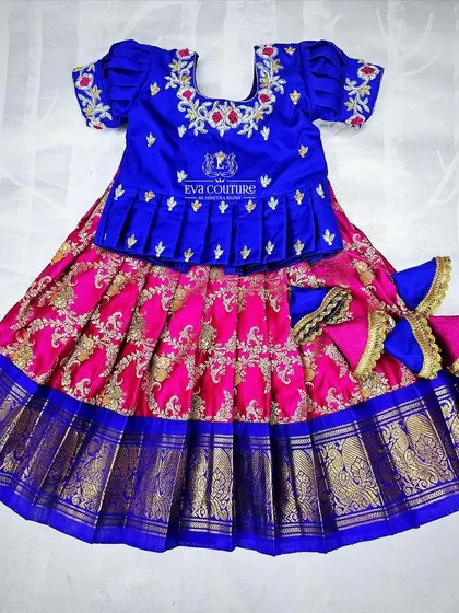 A vibrant pattu lehenga for a 6-month-old, available for customization. The royal blue peplum top is paired with a pink Banarasi skirt and oversized tassels.