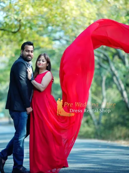 A couple shares a moment outdoors. The woman's simple red gown with a flowing trail is paired with the man's casual blazer and jeans, showing a versatile and relaxed pre-wedding style.