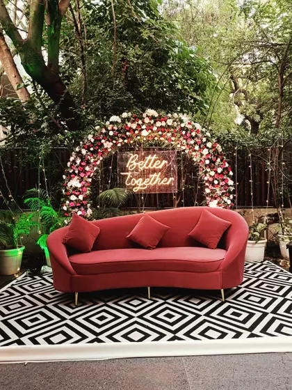 A chic photo booth with a red sofa, a black-and-white geometric rug, and a floral arch with a 'Better Together' neon sign.