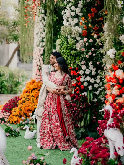 Punya and Teju amidst a spectacular floral backdrop. Her makeup is fresh and radiant, complementing the vibrant colors of the decor and her beautiful red lehenga.