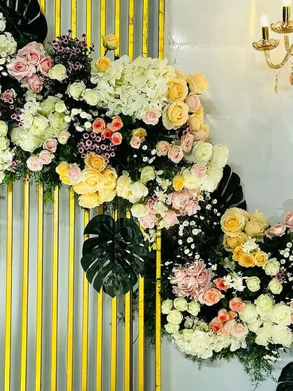 A lush floral arrangement cascading down a modern, golden-paneled wall. The mix of pastel roses, hydrangeas, and tropical monstera leaves creates a luxurious and contemporary feel.