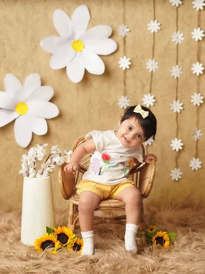 A cheerful baby girl in a daisy-themed setup. Her bright smile and the sunny yellow shorts create a happy and vibrant milestone photo.