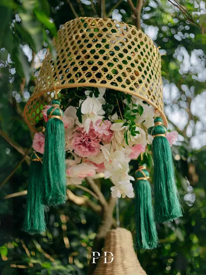 A beautiful decor detail for a boho Mehendi: a rattan basket filled with flowers and adorned with green tassels, hanging from a tree.