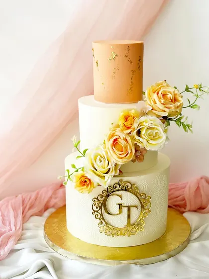 A sophisticated three-tier cake with a custom gold monogram. The design combines a smooth white base, a textured bottom tier, and a warm peach top tier, all brought together with a cascade of yellow and cream roses.