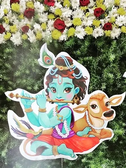 A cute cutout of Lord Krishna with a calf, used as a decorative element against a backdrop of fresh flowers for a naming ceremony.