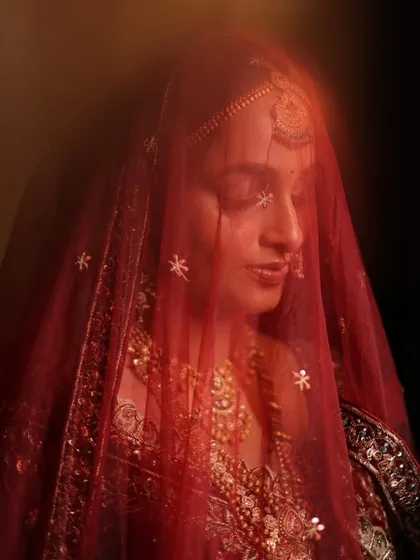 A dreamy, artistic shot of Laxmi through her red veil. This image perfectly captures the soft, ethereal quality of her traditional bridal makeup, highlighting a timeless and graceful aesthetic.